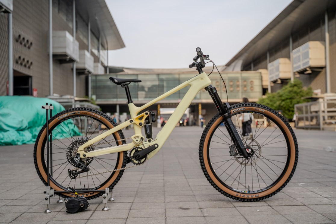 A side view of a lightweight carbon mountain bike with a yellow frame and black front fork, displayed outdoors with an exhibition center in the background.