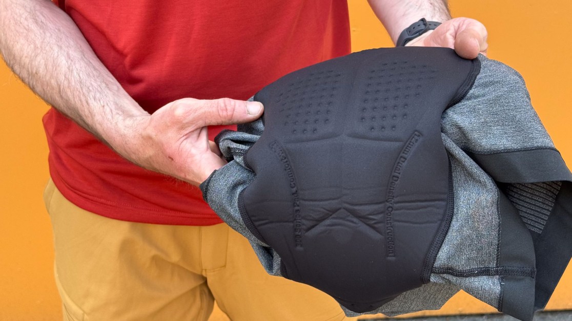 A person holding a padded garment, showcasing its protective features, against a bright orange background.