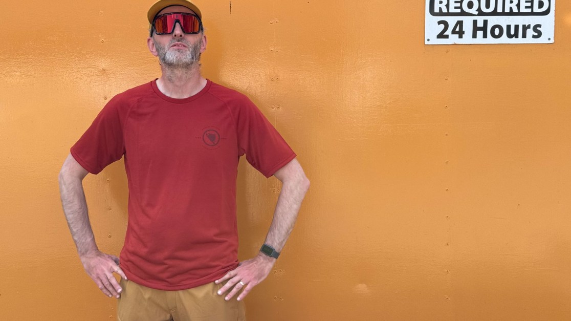A person standing confidently in front of an orange wall, wearing a red shirt and a yellow cap with large sunglasses.