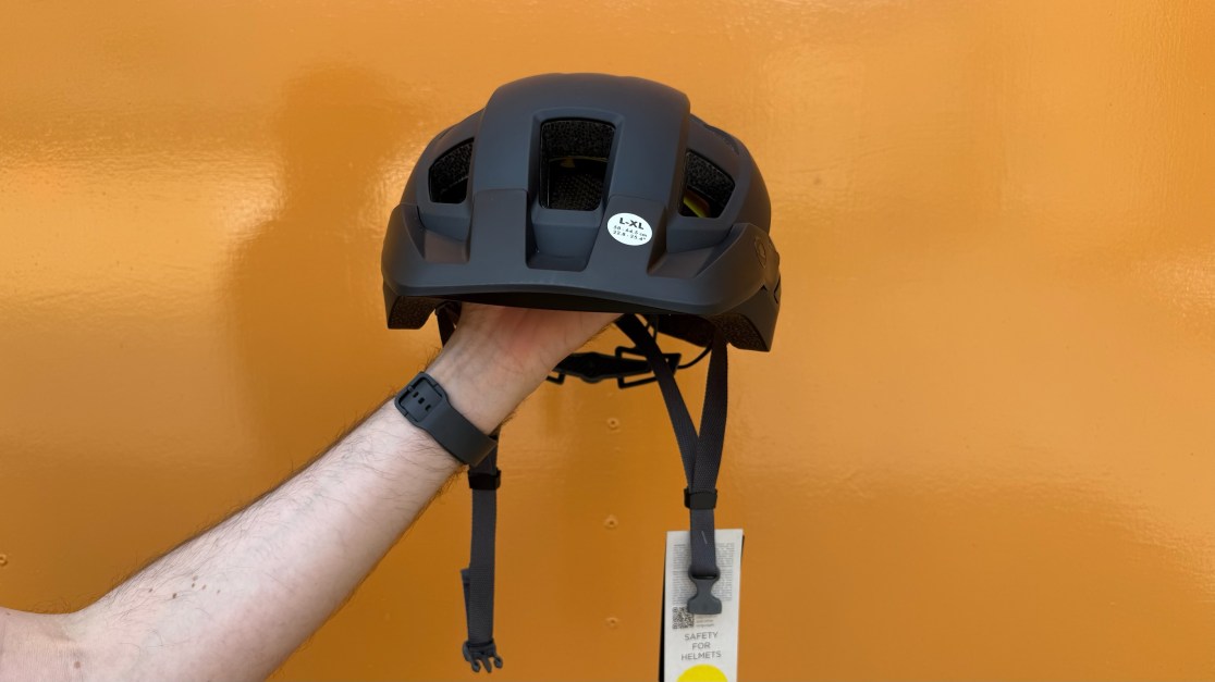 A person holding a black cycling helmet with ventilation holes against an orange background.