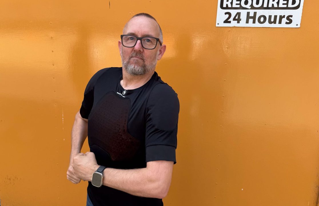 A person wearing a black protective shirt with an intricate design and dark padding stands confidently in front of an orange wall displaying a sign that says 'REQUIRED 24 Hours'.
